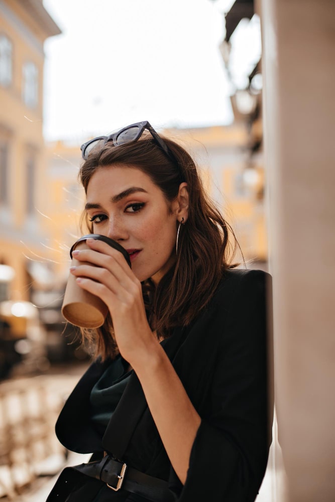 elegant-young-lady-with-brunette-wavy-hair-modern-makeup-black-jacket-drinking-cup-coffee-city-cafe-terrace-looking-straight-min