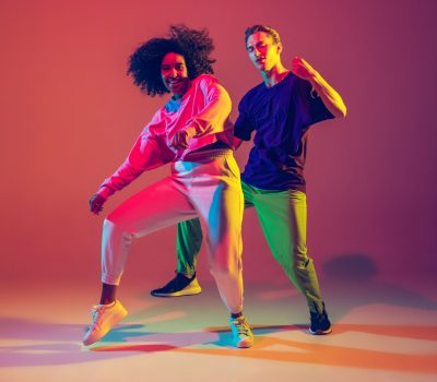 Dance time. Stylish men and woman dancing hip-hop in bright clothes on green background at dance hall in neon light. Youth culture, movement, style and fashion, action. Fashionable portrait.